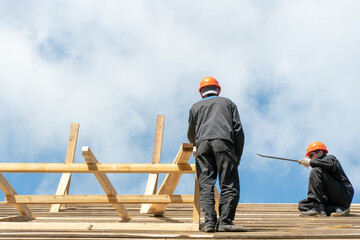 Repair of a wooden roof outdoors on a summer day against the background of blue sky and clouds. A carpenter in special clothes and with a tool installs beams and wood boards.