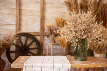 In green glass vase, there was bouquet of dried flowers on wooden bench near backdrop of rural style