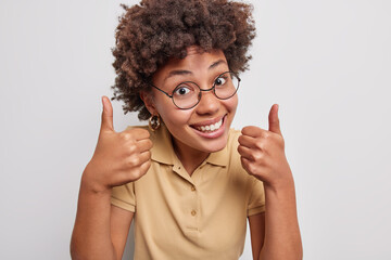 Wall Mural - Pleasant looking curly haired woman smiles happily keeps thumbs up approves something says very good has cheerful expression poses against white background praises you makes compliment. Great deal