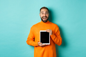 Wall Mural - Technology. Excited adult man in orange sweater showing digital tablet screen, staring amazed at camera, standing over turquoise background