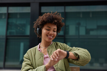 Wall Mural - Horizontal shot of good looking Afro American woman with curly hair checks time on smartwatch listens audio track from social media enjoys leisure pastime dressed in anorak. Millennial hipster