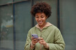 © WHstudio Leushin N - Positive Afro American woman checks newsfeed on mobile phone looks at table of city traffic online uses application or map to find way in big metropolian city wears jacket and hoodie poses outdoor