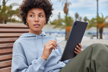 pensive female designer holds tablet with stylus uses public internet connection for creating media 