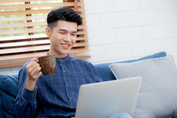 Wall Mural - Young asian businessman smile and work from home with laptop computer online on sofa in living room, freelance business man using notebook and drink coffee on couch, new normal, lifestyle concept.