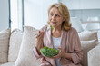 © Grustock - Beautiful elderly woman eats her vegetable salad sitting at home on the couch