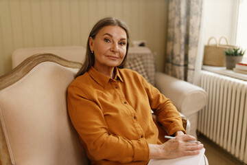 Leisure, rest, hobby and retirement concept. Portrait of attractive serious elderly housewife with gray hair sitting at home, making list of groceries before shopping or writing down her thoughts