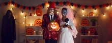 Halloween Banner With Portrait Of Dead Wedding Couple. Happy Groom In Suit Costume Together With Beloved Bride In Romantic White Dress Standing In Dark Room Decorated With Lights And Orange Pumpkins