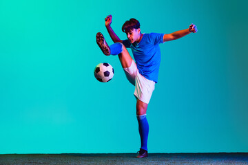 Wall Mural - Horizontal full-length portrait of young sportsman, soccer football player in motion kicking a ball isolated over blue background