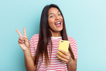 Young Venezuelan woman holding mobile phone isolated on blue background joyful and carefree showing a peace symbol with fingers.