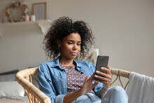 African American Woman Using Smartphone, Relaxing Sitting In Cozy Armchair, Attractive Young Female Looking At Phone Screen, Typing Writing Message In Social Network, Browsing Apps, Shopping Online