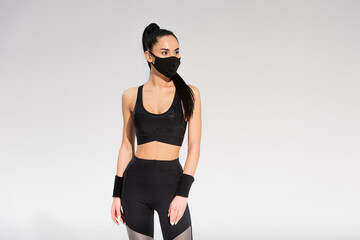 Wall Mural - young sportswoman in sportswear and black protective mask standing on grey