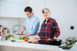 © Alex - pareja joven sonriente cocinando verduras pimiento verde, tomate y calabacin, en una cocina blanca con placa de induccion, utilizan un bol rosa, una sarten de hierro y usan herramientas de madera