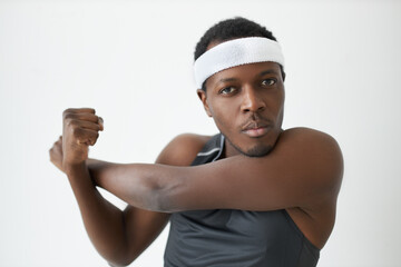 Afrcian American male in fitness headband warming up stretching arms before intensive workout, looking serious and concentrated, focused on body feelings. People, sport, active lifestyle