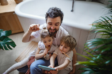 Wall Mural - High angle view of father with two small children brushing teeth indoors at home and taking selfie, sustainable lifestyle concept.