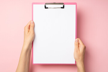 First person top view photo of hands holding pink clipboard with paper sheet on isolated pastel pink background with blank space