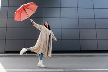 Happy Woman In Beige Coat And White Boots Posing With Red Umbrella In Raised Hand