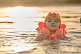 Fototapeta  - A little girl bathes in the water in the sunset light. Child playing on the beach during summer vacation. The child swims in a life jacket and goggles. Safety on the water.