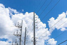 Numerous Tall Metal Power Poles With Electrical Cable Lines And Pale Green Glass Insulators Against A Bright Blue Sky With White Clouds, Horizontal Aspect