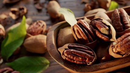 Wall Mural - Pecans in a wooden bowl with pecan leaves and nut shells - close-up video, camera slow motion. The hand takes one of the pecan nut.