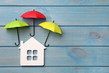 Small Umbrellas And House Figure On Light Blue Wooden Background, Flat Lay. Space For Text