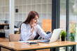 © amnaj - Image of young Asian businesswoman holding a pen using a laptop at a desk with equipment put at the office