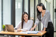 © amnaj - Two young Asian businesswoman work together analyze and suggest pointing a graph using a laptop keyboard at the office.