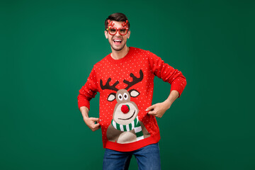 Wall Mural - Portrait of attractive cheerful guy having fun showing deer jumper winter festive isolated over green color background