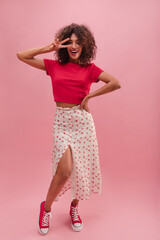 Full shot portrait of stunning curly woman in studio showing peace sign. Image of joyful African woman dressed in long white skirt with cherry and red t-shirt and expressing gladness.