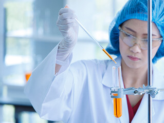 Sticker - Asian female professional scientist in white lab coat safety goggles hygiene cap and rubber gloves holding looking red reagent solution sample in test tube in laboratory full of science equipment