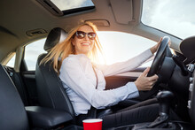 Beautiful Mature Woman With Blond Hair Smiling Sincerely While Driving Car Alone. Business Lady In Sunglasses And Formal Wear Enjoying Her Way To Favorite Work.