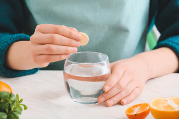 Woman putting ascorbic acid pill in glass of water on table in kitchen