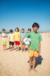 © KAMPUS - Portrait of Asian preteen boy with ball at beach, his friends standing in background. Group of multiethnic kids spending vacation seaside. Summer vacation, friendship concept