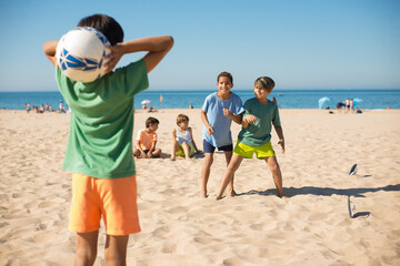 Cheerful boy friends playing football on beach. Preteen boy standing on sand and throwing ball to soccer players. Youth sport, summer vacation concept