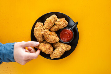 Crispy fried chicken wings with tomato sauce on plate, yellow background. Male hand picking chicken wing, junk food, unhealthy eating concept. Top view copy space