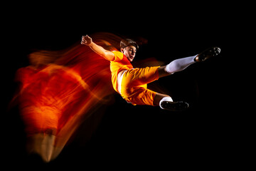Wall Mural - Full-length horizontal portrait of young sportsman, soccer football player training isolated over black background in mixed light
