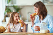 happy loving family, mother and daughter, sitting at table and having breakfast in morning at home. woman and girl eat fresh bread and drink cow's milk, and have nice time together in kitchen