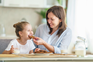 happy loving family, mom and daughter, playing sitting at table and having breakfast at home in morning. woman and girl eat oatmeal cookies and drink cow's milk, and have nice time together in kitchen