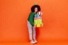 Full Length Portrait Of Woman With Afro Hairstyle Wearing Green Sweater Holding Lots Of Heavy Presents, Looking At Camera With Astonished Expression. Indoor Studio Shot Isolated On Orange Background.