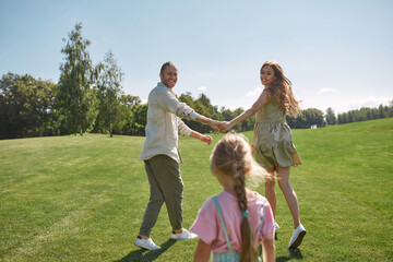 Joyful parents holding hands, looking at their little daughter, having fun together in green park on a summer day