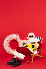 Wall Mural - Santa in sunglasses holding retro boombox and cocktail in coconut on deck chair near swim ring on red background