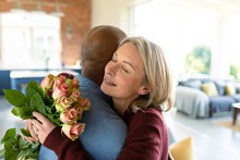Happy Senior Diverse Couple In Living Room Embracing, Holding Flowers