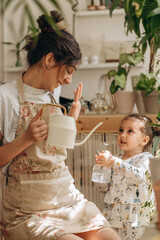 Wall Mural - Mixed race family woman and her little daughter is watering houseplants at home and having fun splashing water.Home gardening.Family leisure,hobby concept.Biophilia design and urban jungle concept