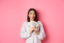 Shopping Concept. Dreamy Asian Woman Thinking, Holding Money Dollars And Looking Aside Thoughtful, Standing Over Pink Background