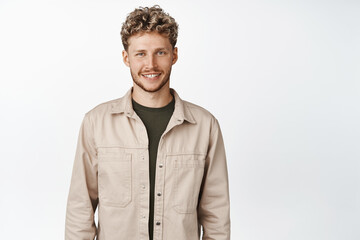 Young handsome man with curly stylish haircut, smiling at camera and looking happy, standing in autumn clothes, white background