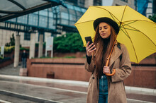Beautiful Woman Using A Smartphone And Holding A Yellow Umbrella While Walking In The City On A Rainy Day