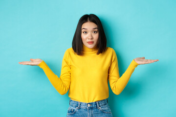 Wall Mural - Image of indecisive asian woman shrugging shoulders, spread hands sideways and looking clueless at camera, standing indecisive against blue background