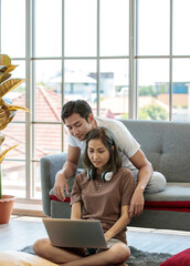 Wall Mural - Young attractive Asian couple sitting and looking at laptop together in living room. Woman sitting on the floor wearing headphones and man lying on couch. Concept for love and couple relationship