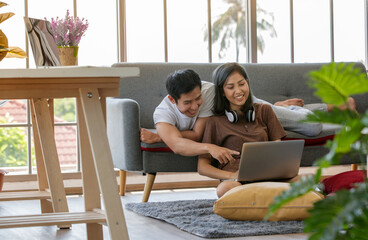 Wall Mural - Young attractive Asian couple sitting and looking at laptop together in living room. Woman sitting on the floor wearing headphones and man lying on couch. Concept for love and couple relationship