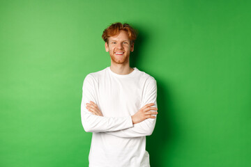 Young ambitious man with red hair standing over green background, holding hands crossed on chest and smiling