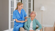 © AboutLife - Young nurse in uniform listens to senior patient while sitting on armrest of comfortable armchair